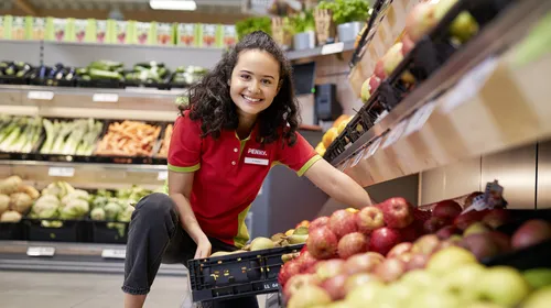 Eine junge PENNY Auszubildende hebt einen Korb mit Obst in ein Regal in einem PENNY Markt