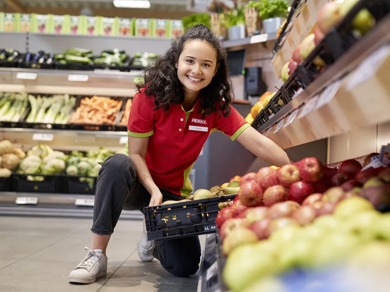 Eine junge PENNY Auszubildende hebt einen Korb mit Obst in ein Regal in einem PENNY Markt