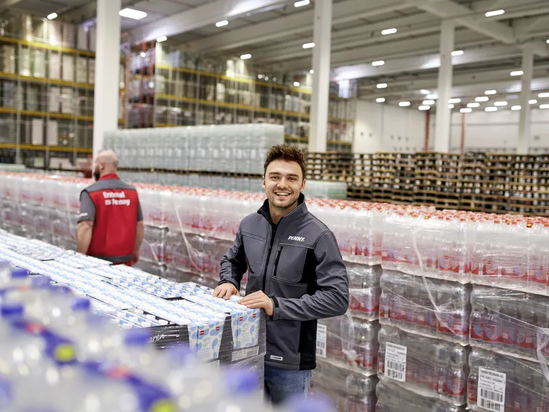 Ein junger PENNY Mitarbeiter steht in einem Lager. Ein weiterer Mitarbeiter läuft im Hintergrund durch einen Gang.