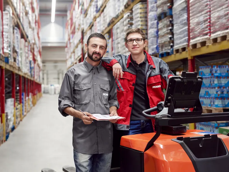 Zwei Mitarbeiter stehen nebeneinander in einem Logistikstandort. Rechts auf dem Bild ist ein Gabelstapler.