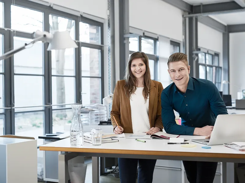 Eine junge Mitarbeiterin und ein junger Mitarbeiter in Bürokleidung in der Zentrale, die vor einem Schreibtisch stehen