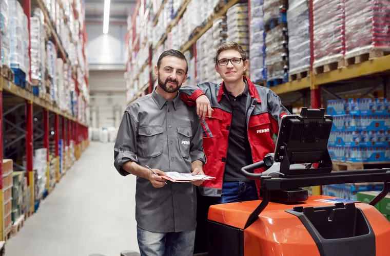 Zwei PENNY Mitarbeiter stehen nebeneinander in einem Logistikstandort. Rechts auf dem Bild ist ein Gabelstapler.